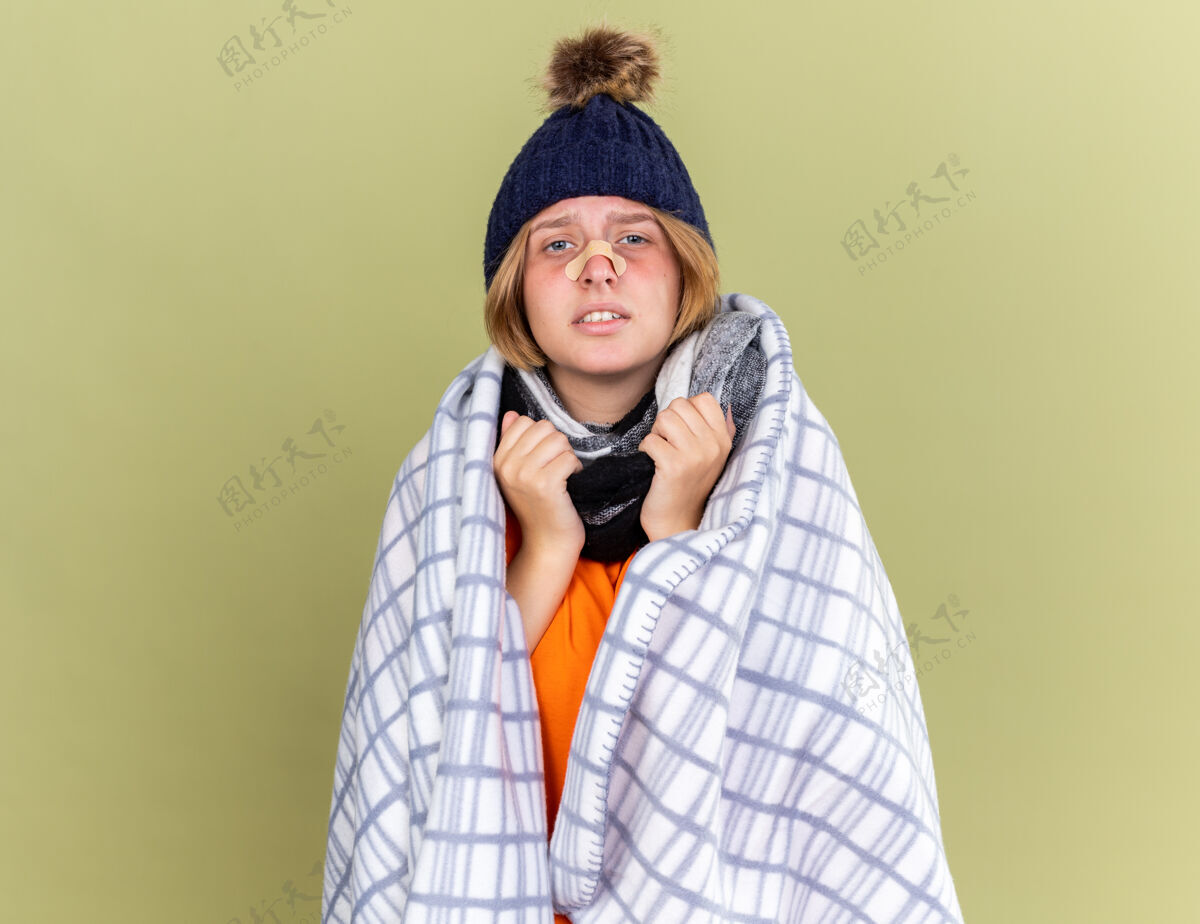 站不健康的年轻女子 戴着暖和的帽子 脖子上围着围巾 裹着毛毯 忍受着感冒的折磨 鼻子上贴着补丁 站在绿色的墙壁上 感觉不舒服补丁感觉不适
