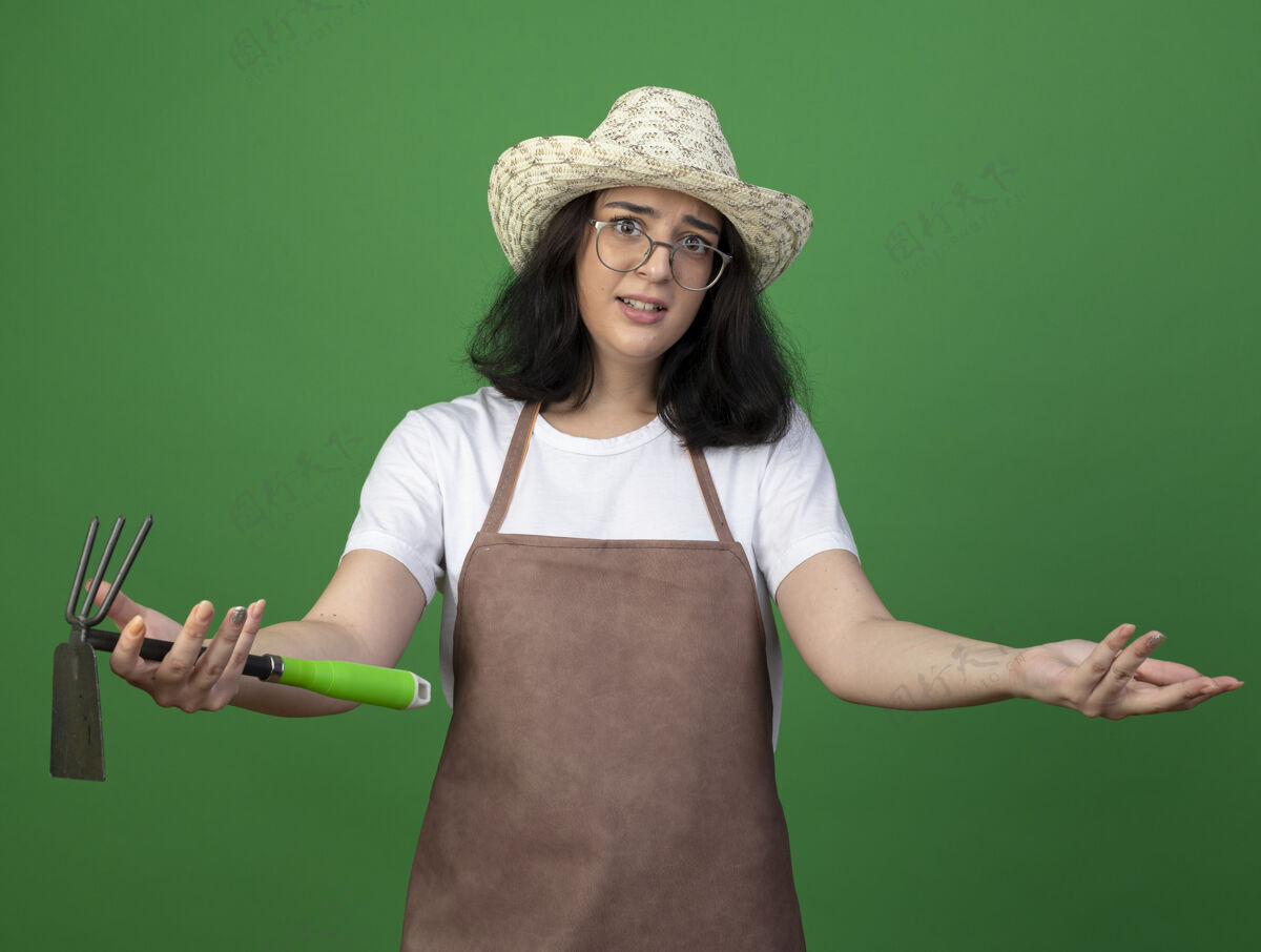 年轻失望的年轻黑发女园丁戴着眼镜和制服 戴着园艺帽 拿着锄头耙子 在绿色的墙上保持手的张开和隔离开放光学姿势