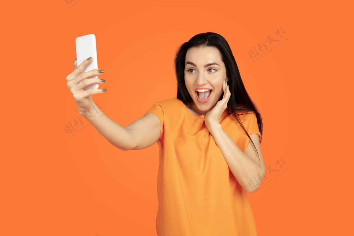 身长橙色工作室背景上的白种人年轻女子肖像穿着衬衫的漂亮深色女性模特人类情感的概念 面部表情 销售 广告广告空间自拍 赌赢女性财务黑发