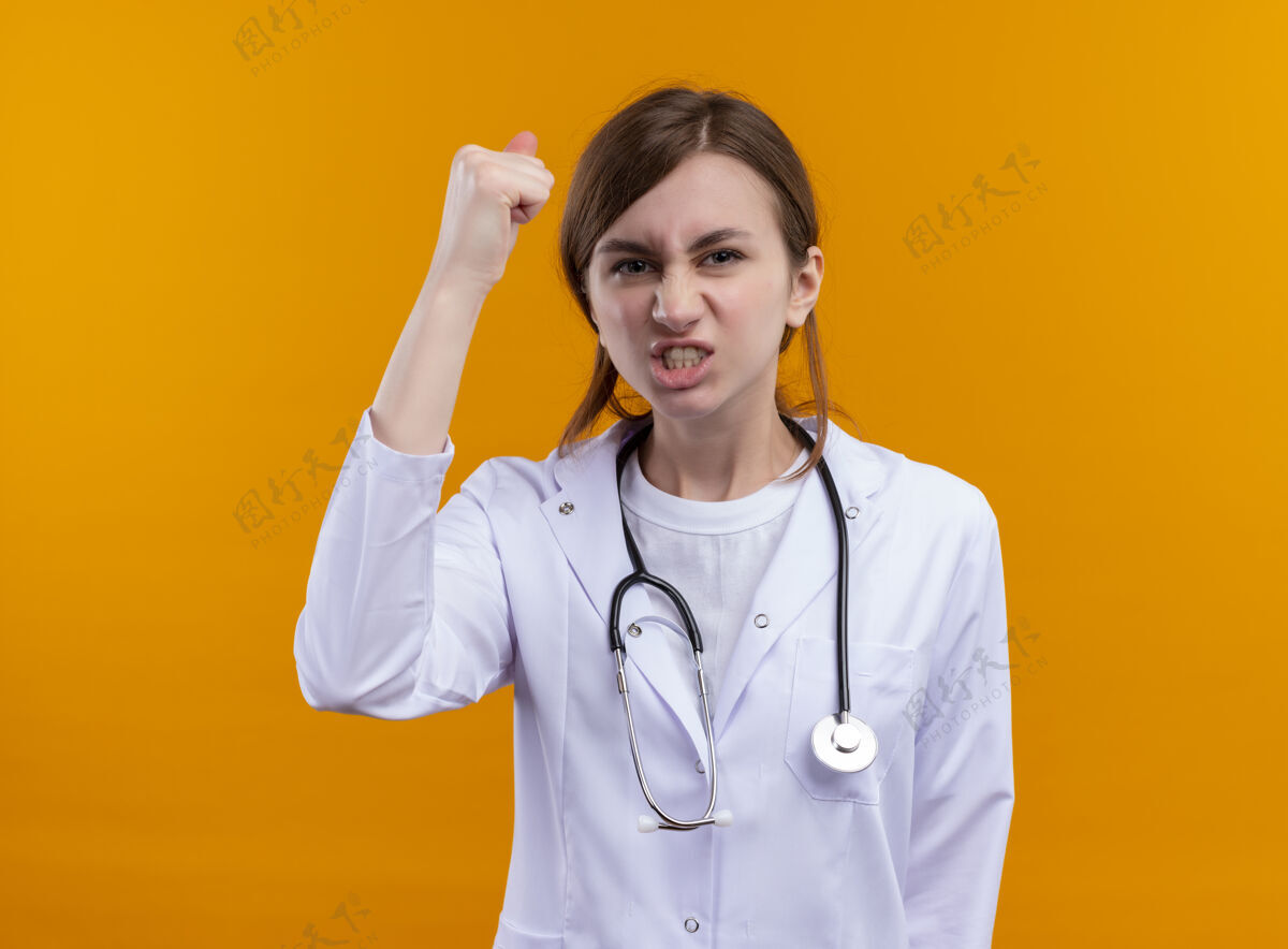 年轻自信的年轻女医生穿着医用长袍 手持听诊器 高举拳头 站在孤立的橙色空间和复印空间女性拳头长袍