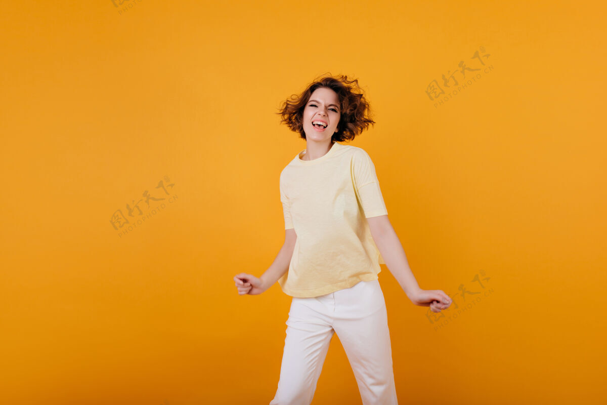 舞蹈穿着黄色t恤的迷人年轻女孩摆出充满感情的姿势穿着白色裤子跳舞的时髦白人女孩的室内肖像 做着滑稽的面孔年轻青少年喜悦