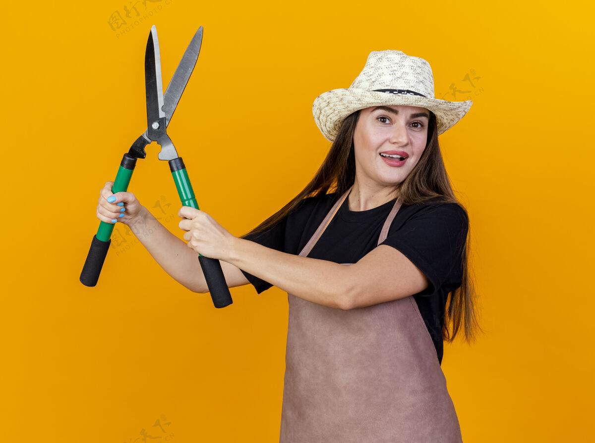 帽子微笑美丽的园丁女孩穿着制服和园艺帽子提出剪子孤立的橙色背景制服提高花园