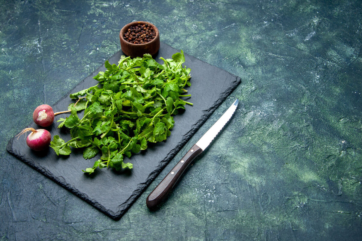 前正面图香菜包新鲜萝卜胡椒在木制砧板和刀的右侧绿色黑色混合色背景与自由空间切壁板蔬菜