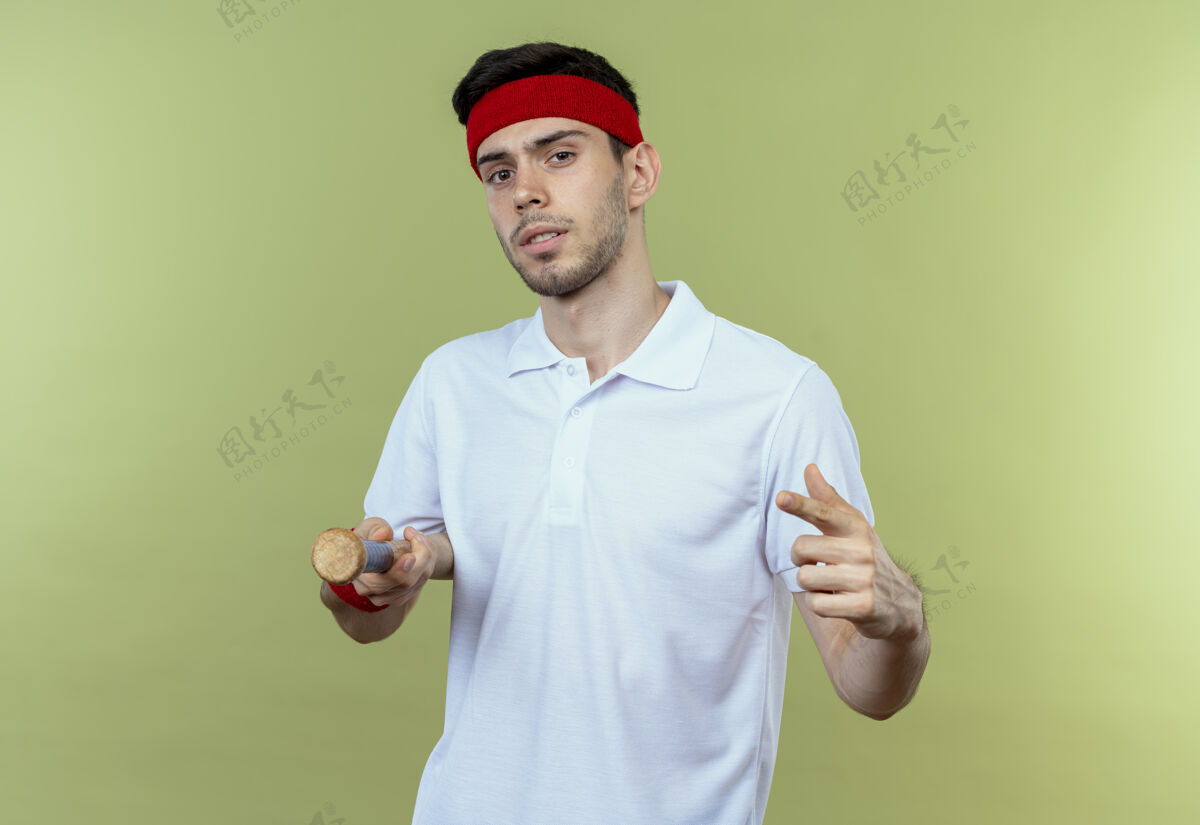 年轻人戴着头巾的年轻运动型男子拿着棒球棒 手指指着塞梅拉 站在绿色的墙上 看上去很自信棒球站蝙蝠