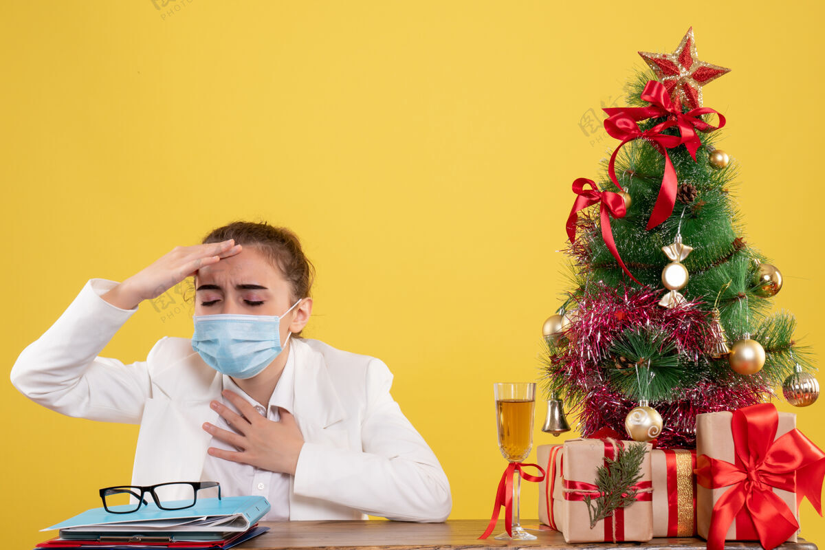 坐着正面图女医生戴着防护面具坐在黄色背景上 戴着圣诞树和礼盒生活背景母亲
