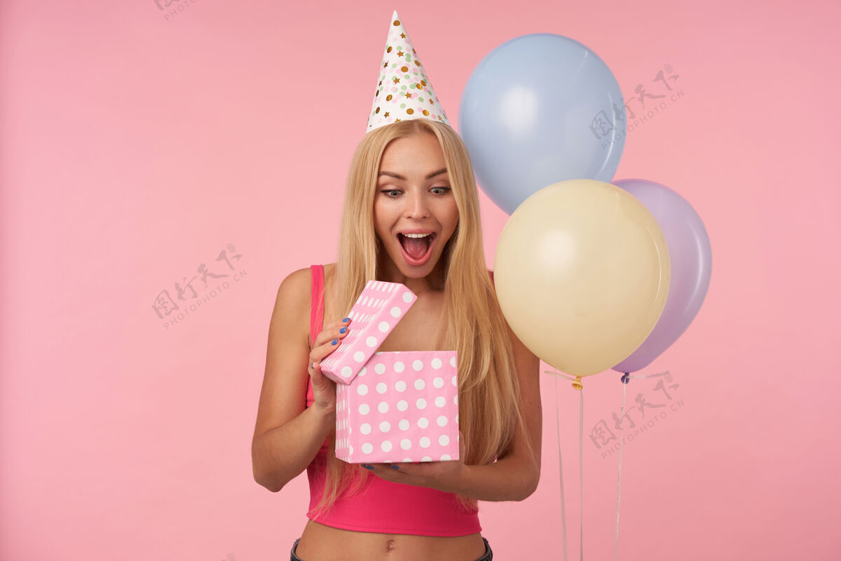 漂亮快乐的金发女郎 留着长长的头发 打开礼物 对自己的内容感到惊讶 在粉色背景下摆出五颜六色的气球 睁大眼睛和嘴巴五彩20多岁休闲