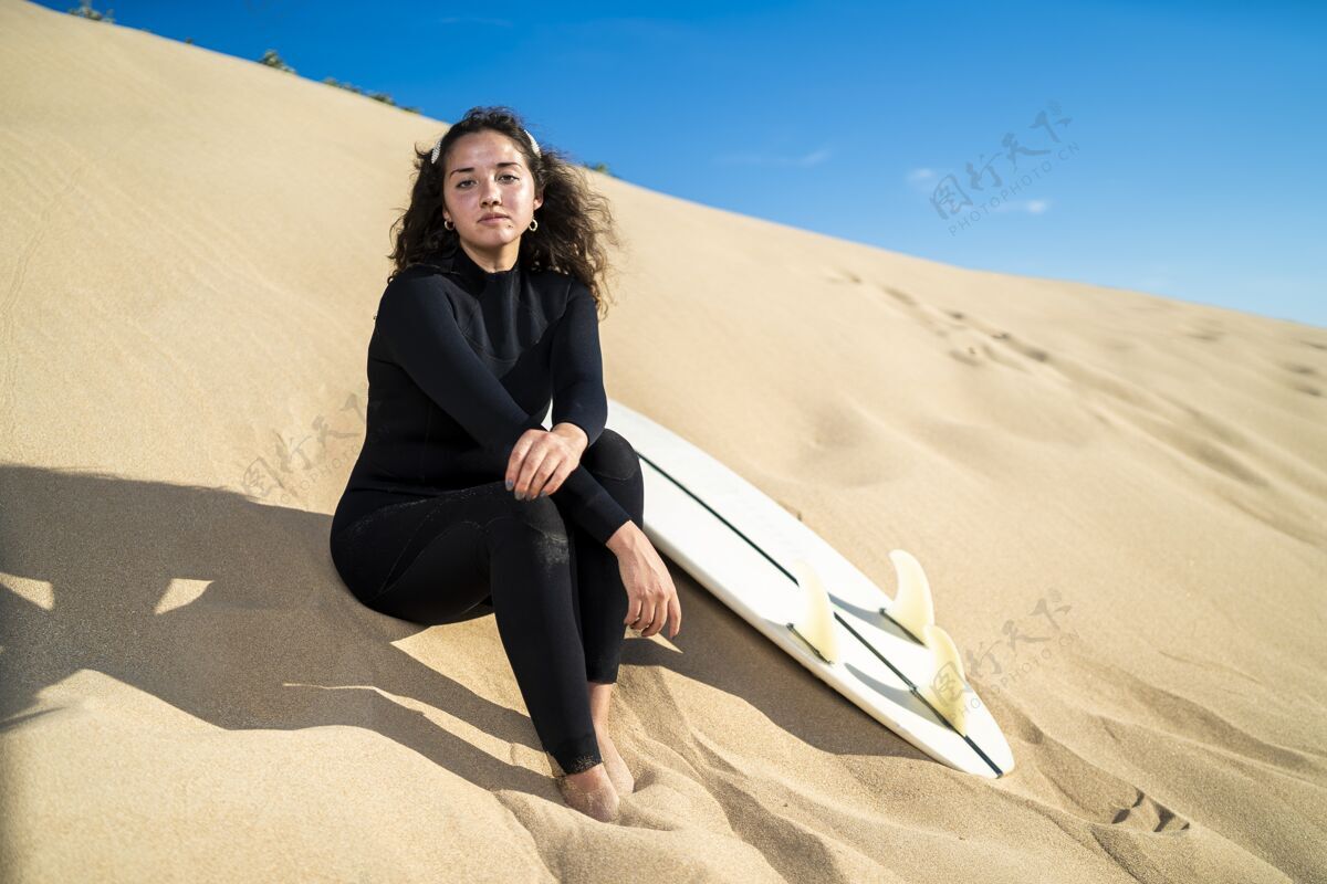 活跃一个迷人的女性坐在沙山与冲浪板在一边的照片休闲海岸海滩