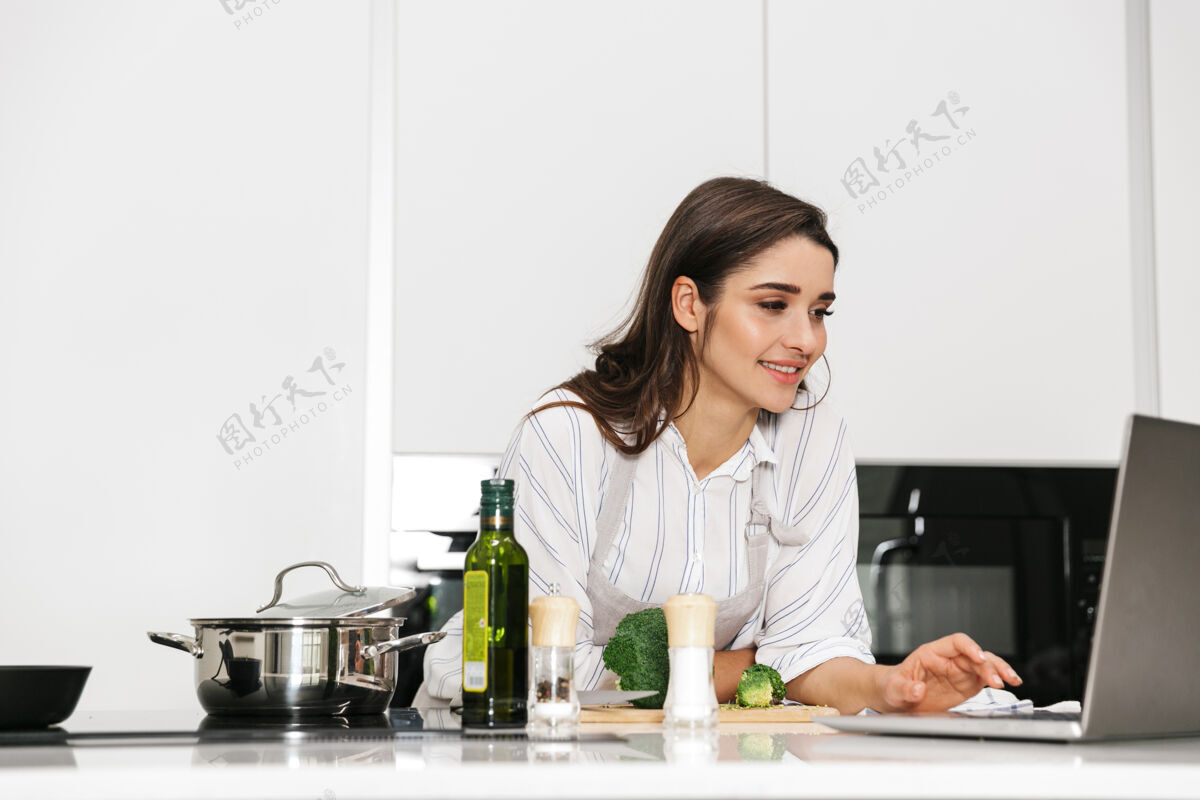 人漂亮的年轻女子在厨房的锅里煮健康的晚餐 看着笔记本电脑室内漂亮食谱