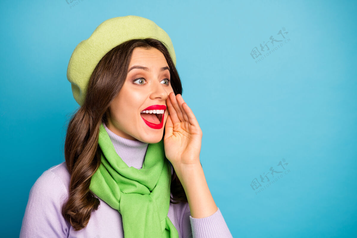 讲话搞笑旅行者女士手拉手近嘴尖叫出售购物秘密信息戴绿色贝雷帽紫色高领围巾隔离蓝色墙壁新闻好奇声音