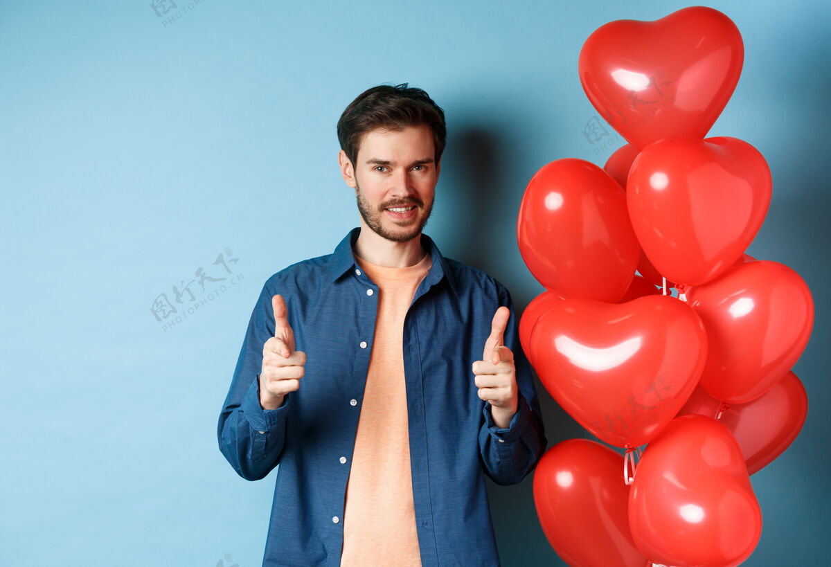 激情情人节快乐自信的男人指着相机微笑着 站在蓝色背景上的气球男人伴侣男朋友