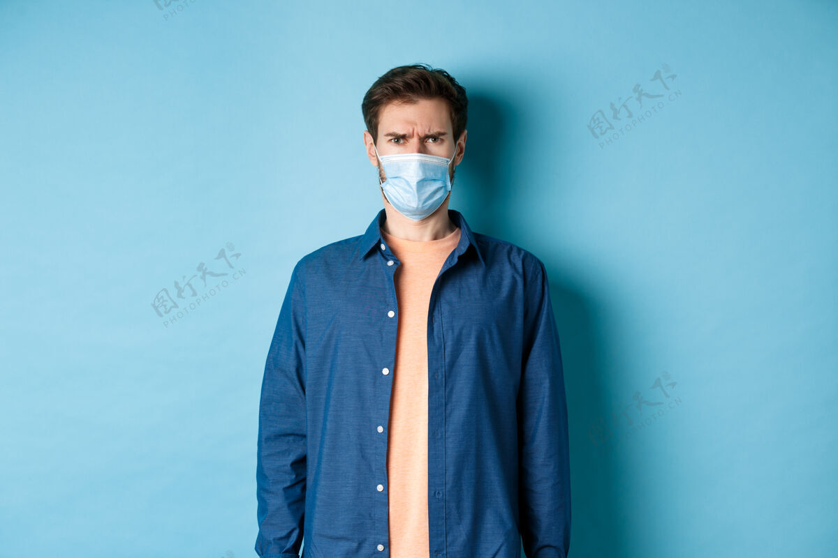 社会距离Covid-19和医疗保健概念戴着医学面具的失望的家伙皱着眉头 看起来很困惑 站在蓝色的背景上男性立场休闲