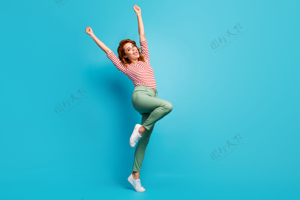 微笑美女疯狂啦啦队队长举起拳头双手支持运动队穿着休闲红白衬衫绿裤子鞋孤立的蓝色冠军年轻女孩