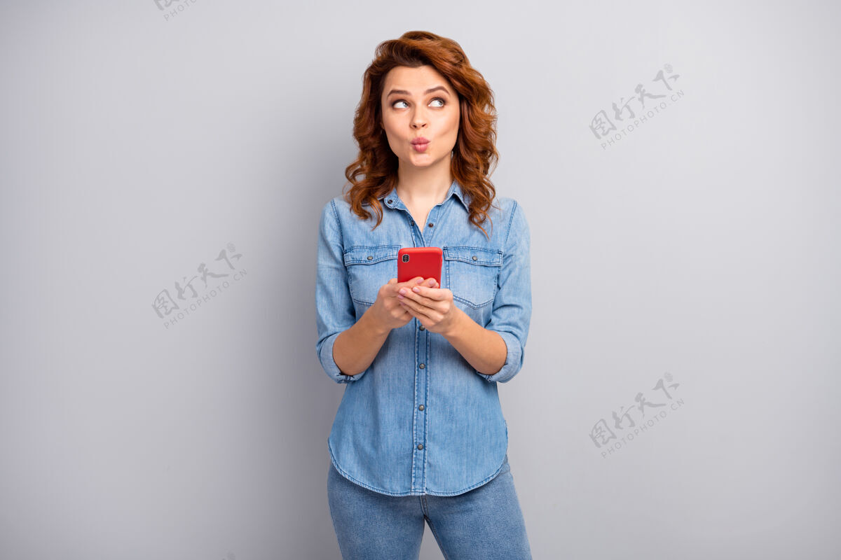 智能浪漫正面开朗的博客写真女人用智能手机看文案空间想思想想文字型博客帖子穿牛仔牛仔裤衬衫隔离灰色墙壁寒冷年轻电话
