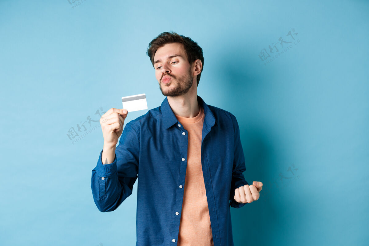 男人有趣的家伙亲吻他的塑料信用卡 看起来很满意 购物打折 站在蓝色背景上年轻购物者买家
