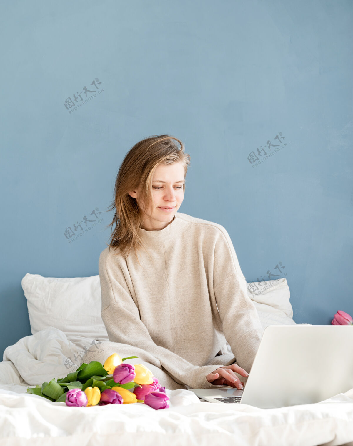 复活节快乐微笑的女人穿着睡衣坐在床上 愉快地赏花 工作在笔记本电脑上惊喜花床