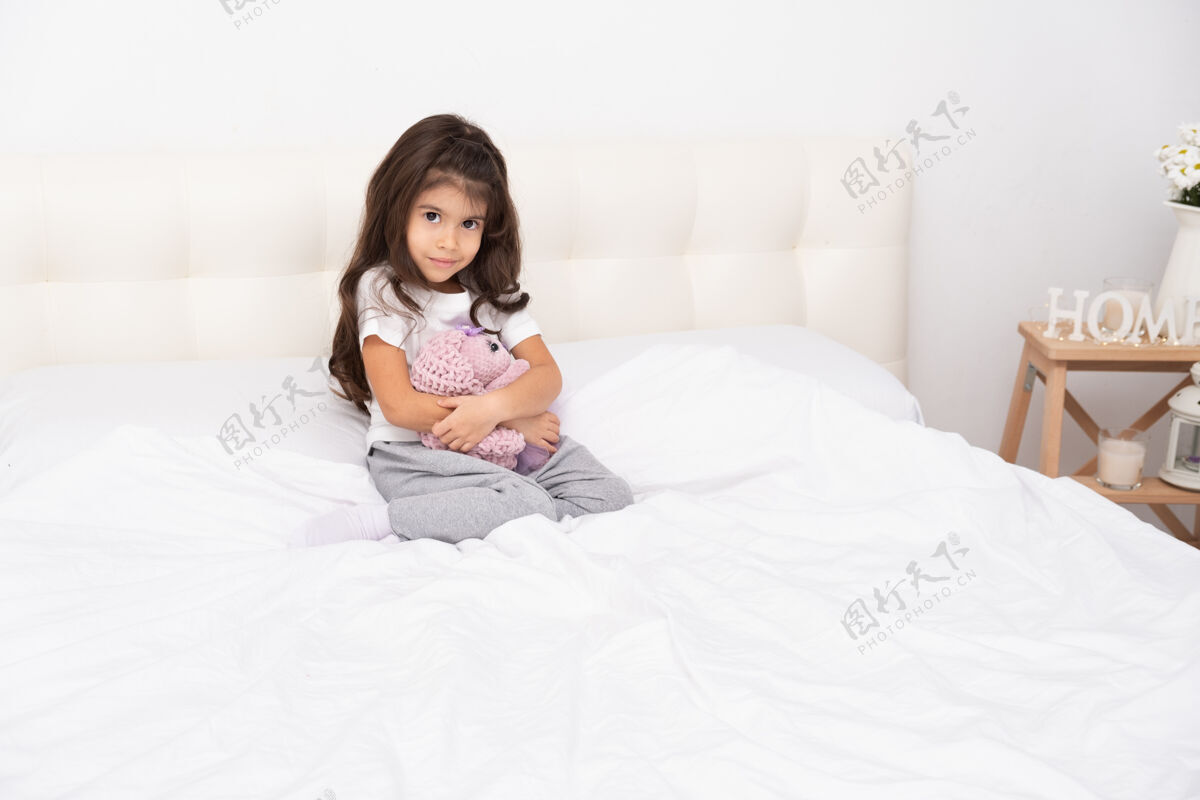 放松穿着家装的小女孩和泰迪兔坐在家里的床上纺织品护理孩子
