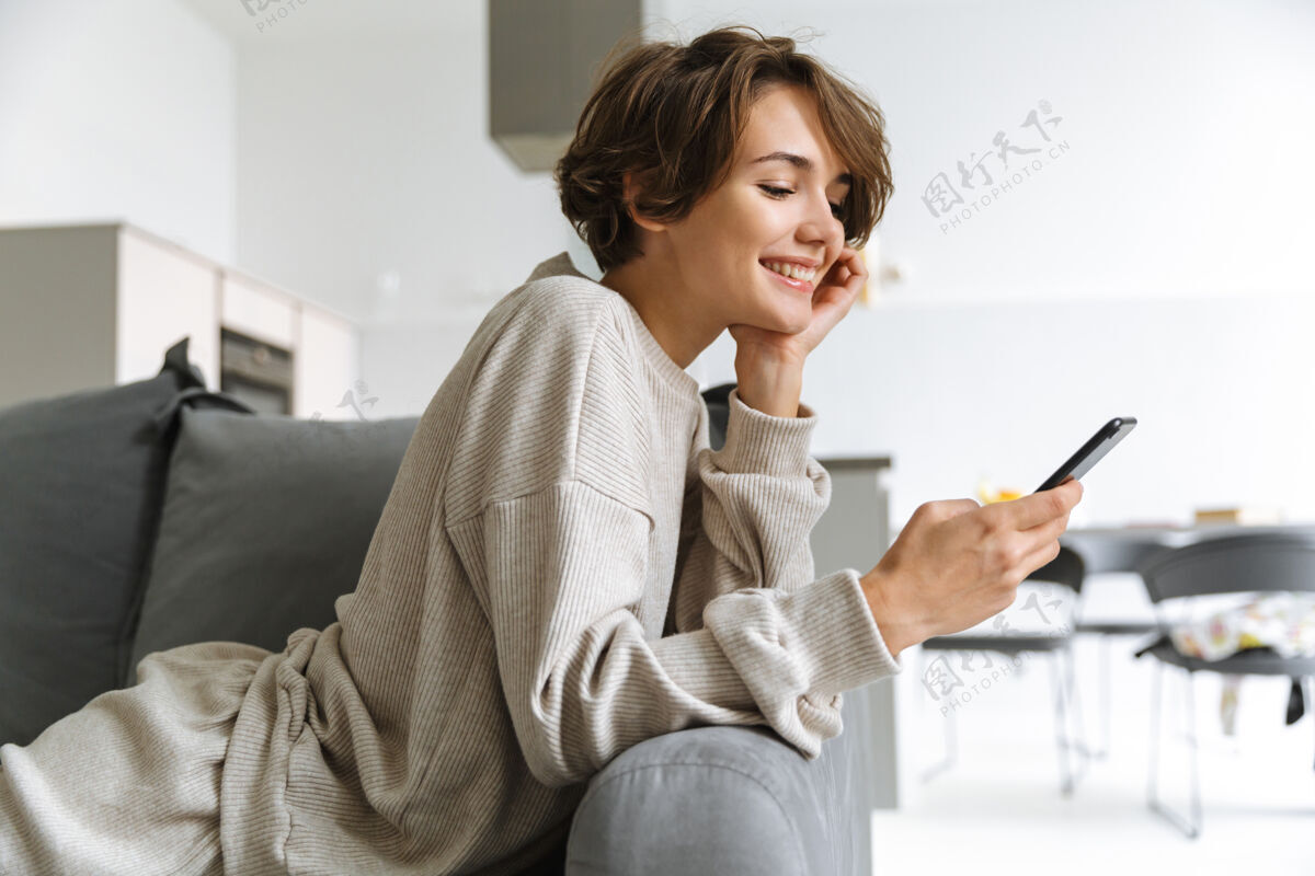 短信快乐的年轻女子坐在家里的沙发上 用手机年轻购物电话
