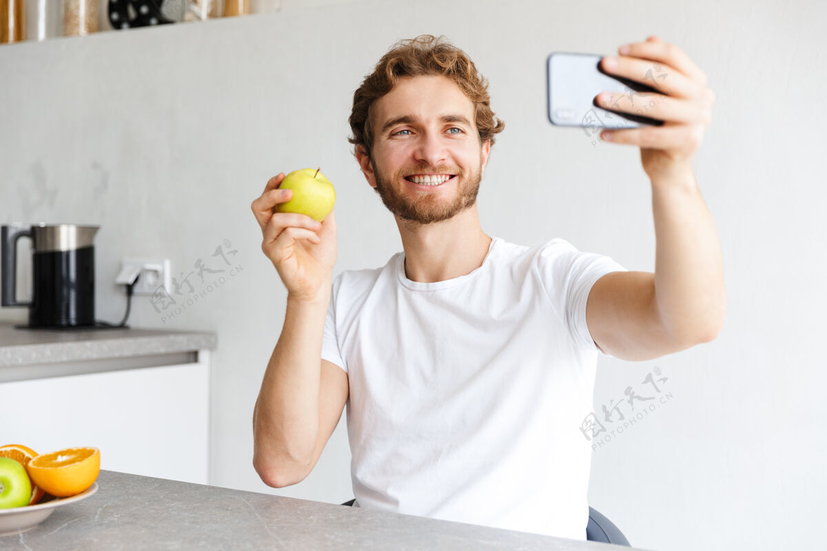 软图为一位快乐的留着胡子的年轻人在家里的餐桌上用水果通过电话自拍室内放松白种人
