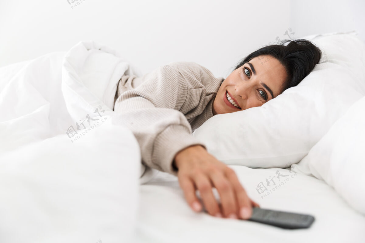 小玩意图为30多岁的黑发女子在家中穿着白色亚麻布躺在床上使用智能手机吸引聊天白种人