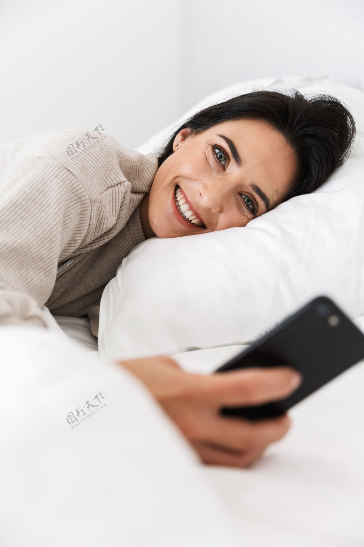 沙发30多岁的快乐女人在家里穿着白色亚麻布躺在床上使用智能手机的照片女人舒适肖像