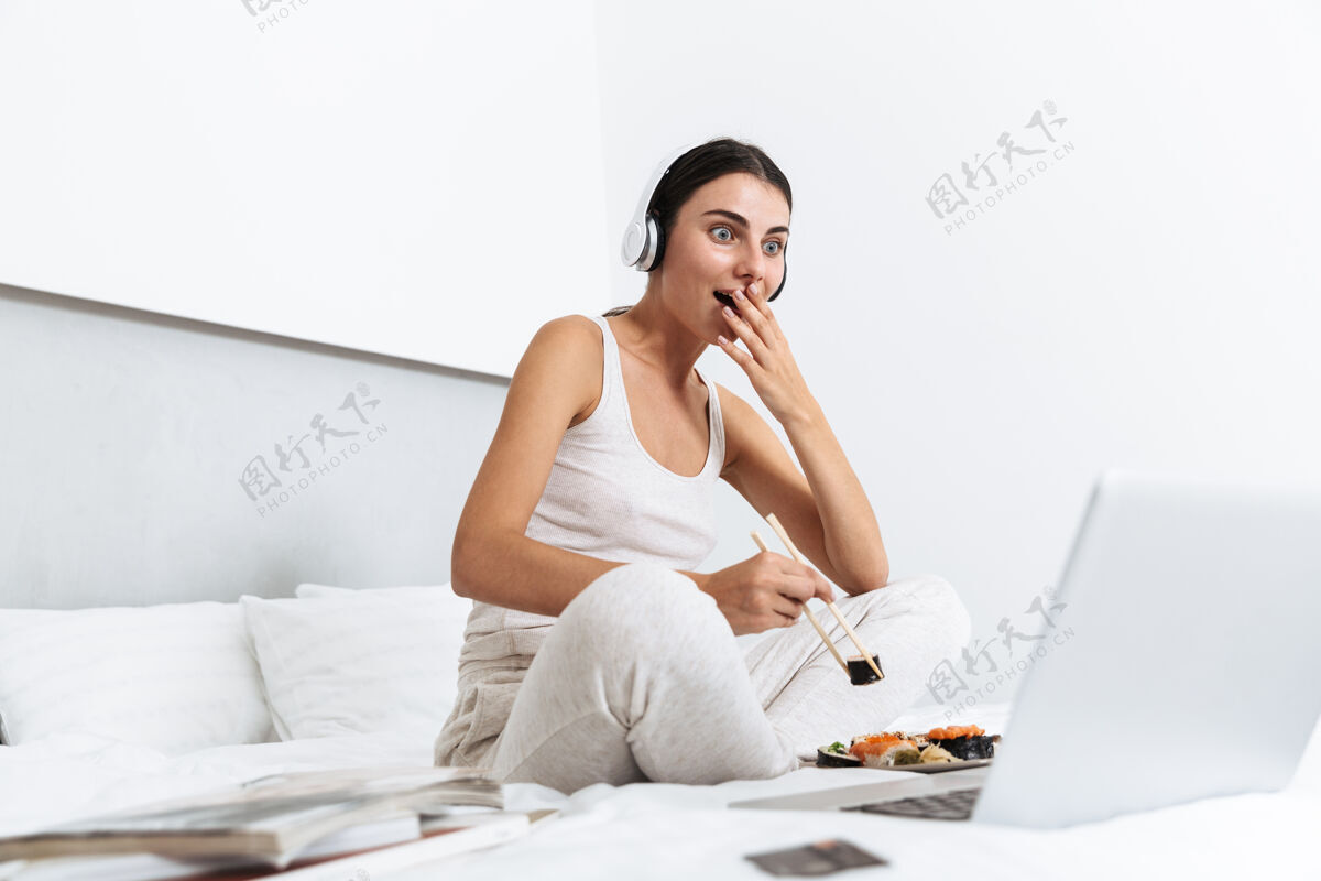 享受美丽的年轻女子在家里的床上放松 戴着耳机听音乐 吃着盘子里的寿司笔记本电脑听力新鲜