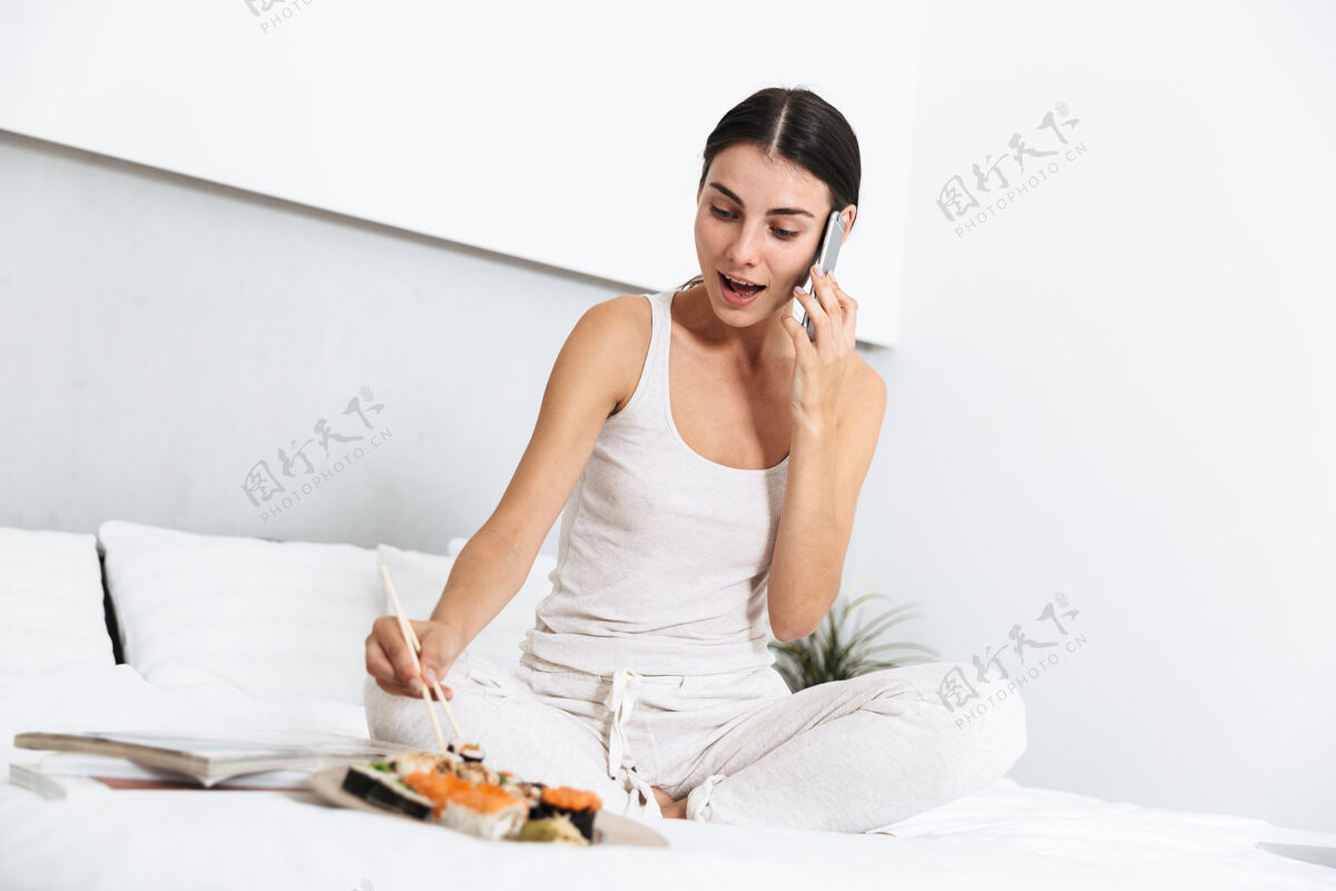 听力美丽的年轻女子在家里的床上放松 吃着盘子里的寿司 讲着手机年轻午餐连接