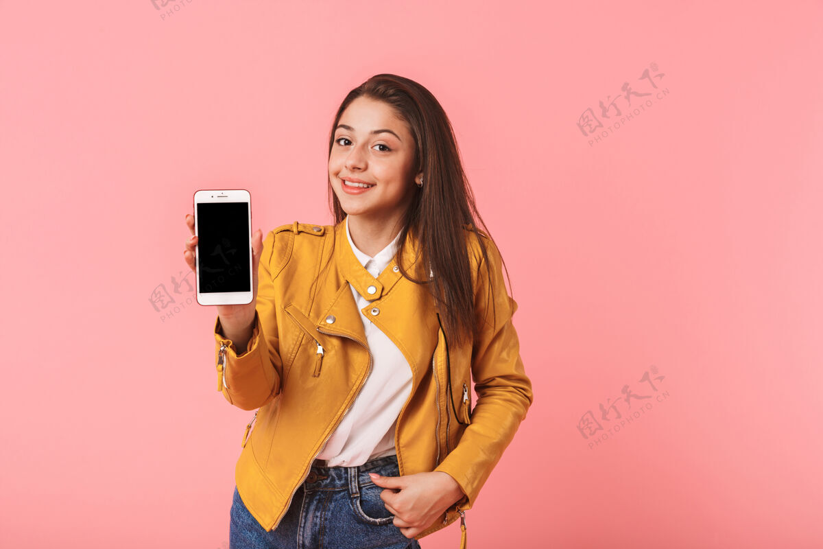 微笑快乐的年轻女子孤零零地站在粉红色的墙上 展示着黑屏手机成人站立手机