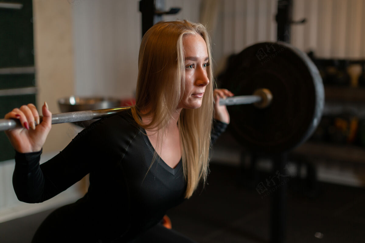 年轻女人一个强壮的金发女运动员背着杠铃蹲着 在体育馆里向前看运动有氧肌肉