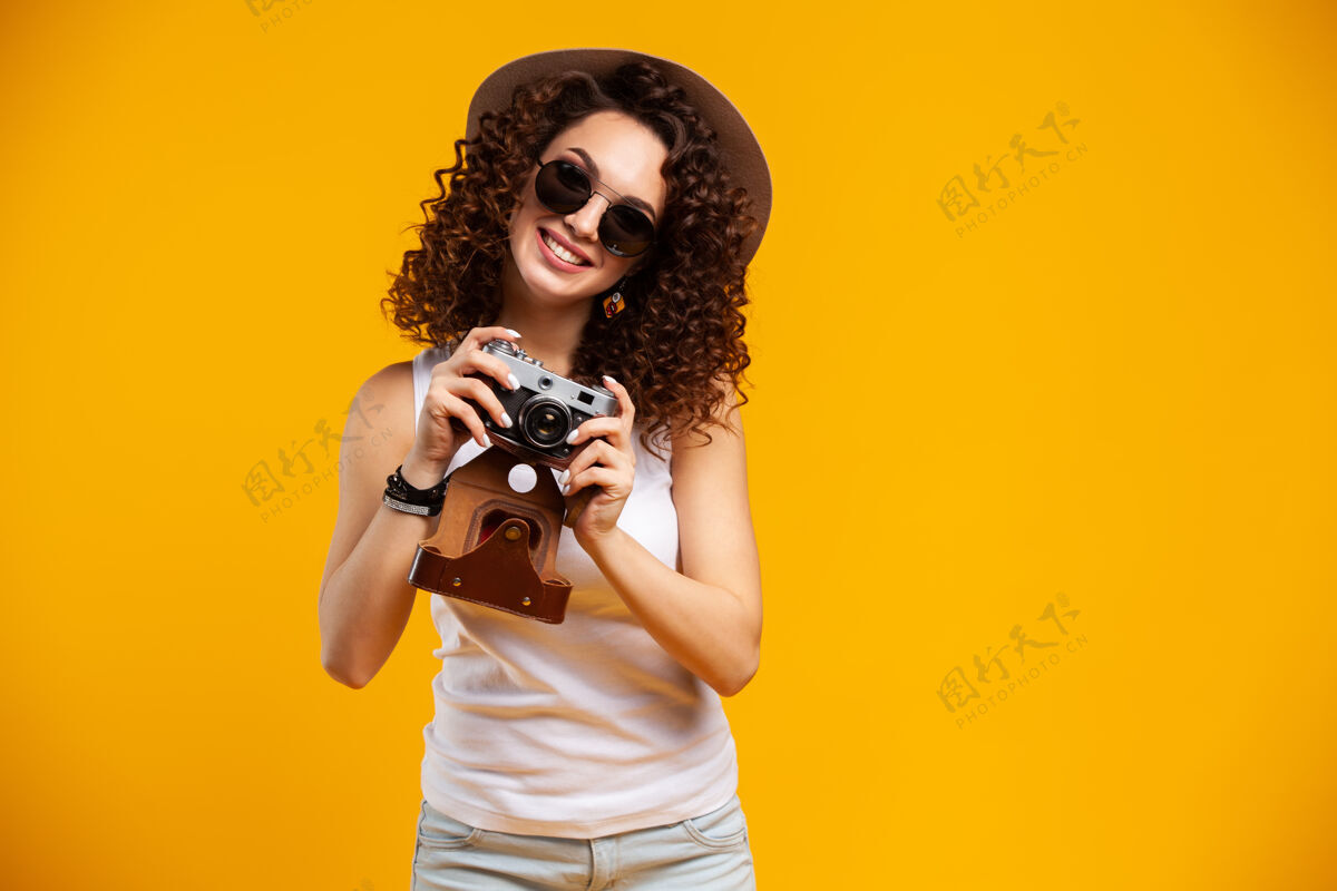 年轻戴着眼镜的笑女青年在复古老式照相机上拍照的肖像 隔离在明亮的黄色上微笑惊人姿势