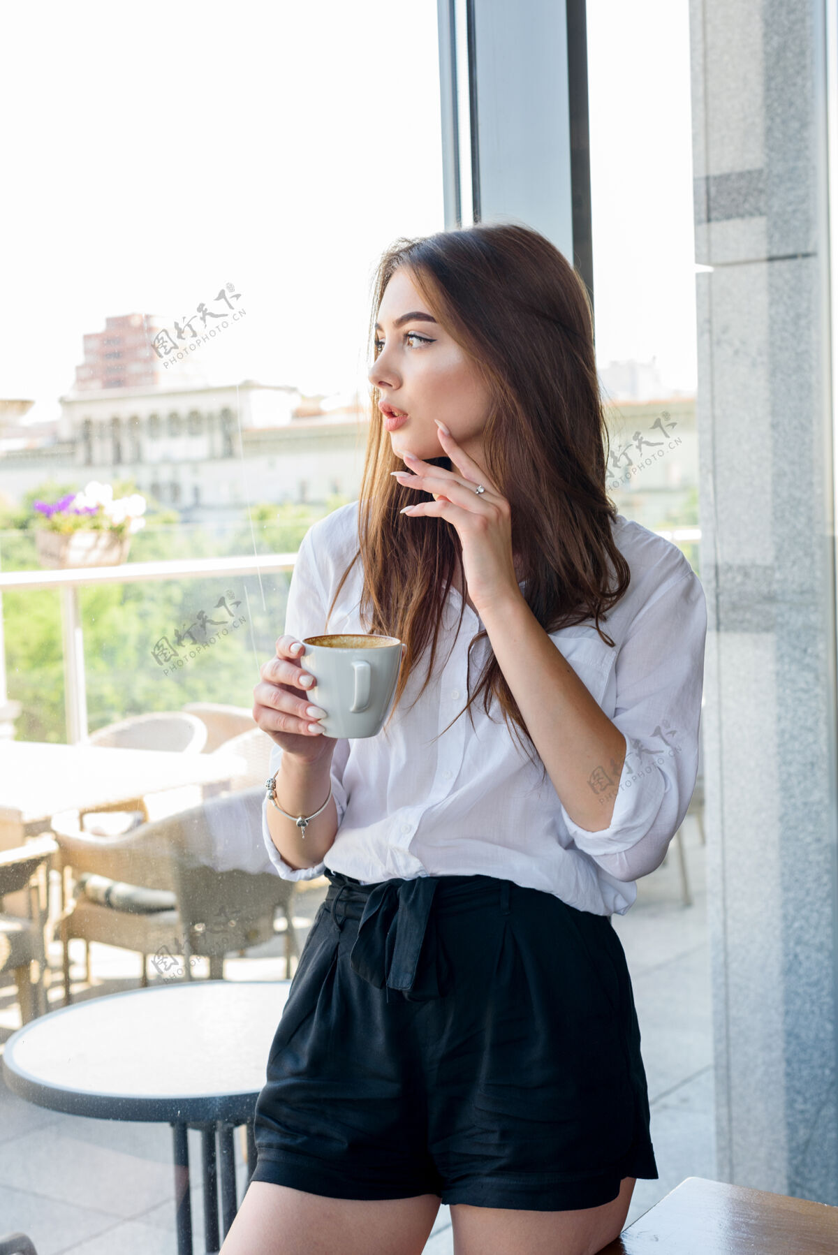 咖啡馆一个年轻漂亮的棕色头发的女孩 穿着白衬衫 在靠窗的咖啡馆里喝着卡布奇诺早餐年轻漂亮