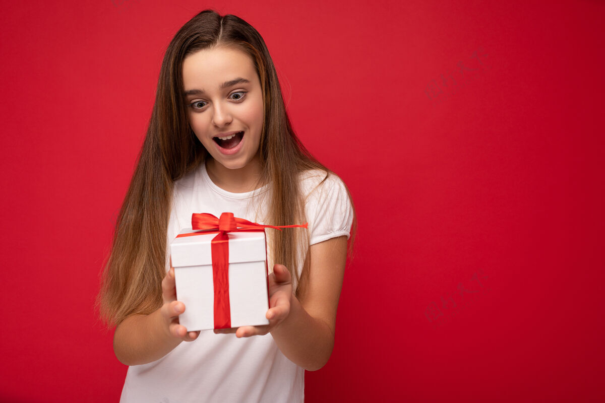 礼物照片中美丽的积极快乐的小女孩儿被隔离在红色的地面上 穿着休闲的白色t恤 墙上拿着礼盒 正在吃饭有趣空虚空间孩子礼品盒盒子