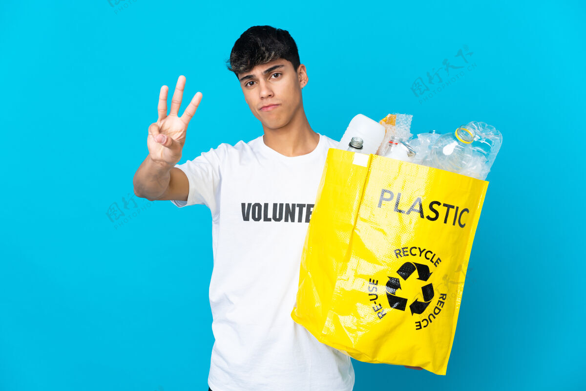志愿者拿着装满塑料瓶的袋子在蓝色的墙上快乐地回收 用手指数着三个减肥垃圾回收