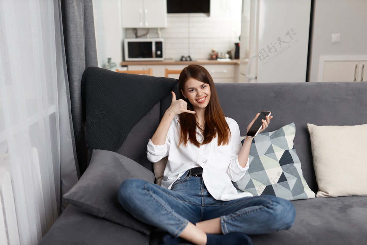 博客快乐的女人坐在公寓的沙发上打电话使用快乐便携式信息设备
