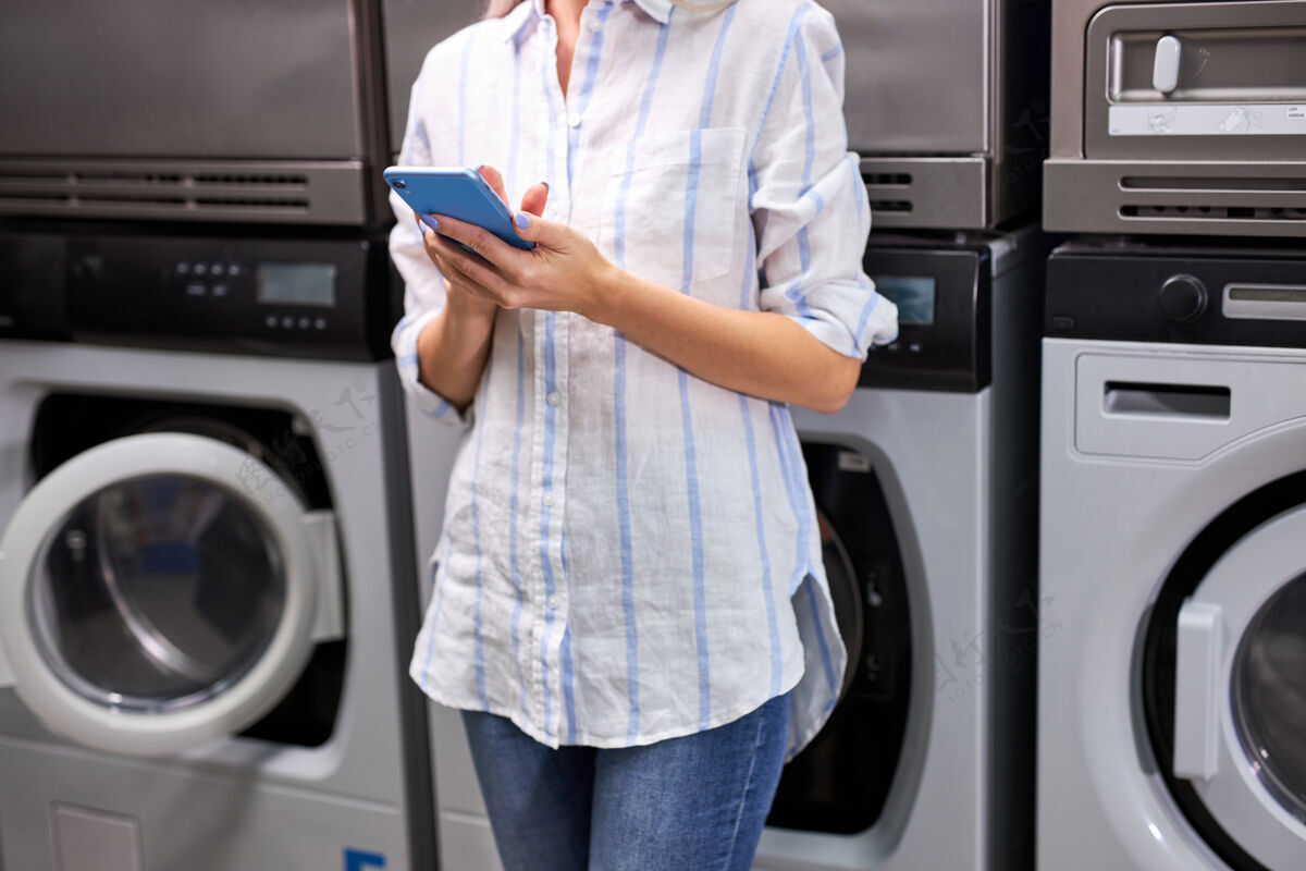 洗衣机一个女人用手机站在洗衣机旁边 等着洗衣机的尽头洗涤.不规则剪裁洗衣房里不可辨认的女人电器人坐着