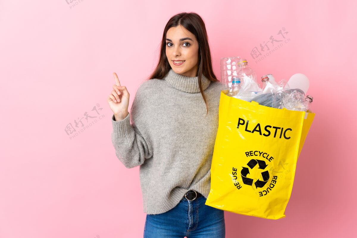 保护年轻的高加索女子拿着一个装满塑料瓶的袋子 在粉红色的背景上孤立地指着旁边赠送的一款产品方向信心女性