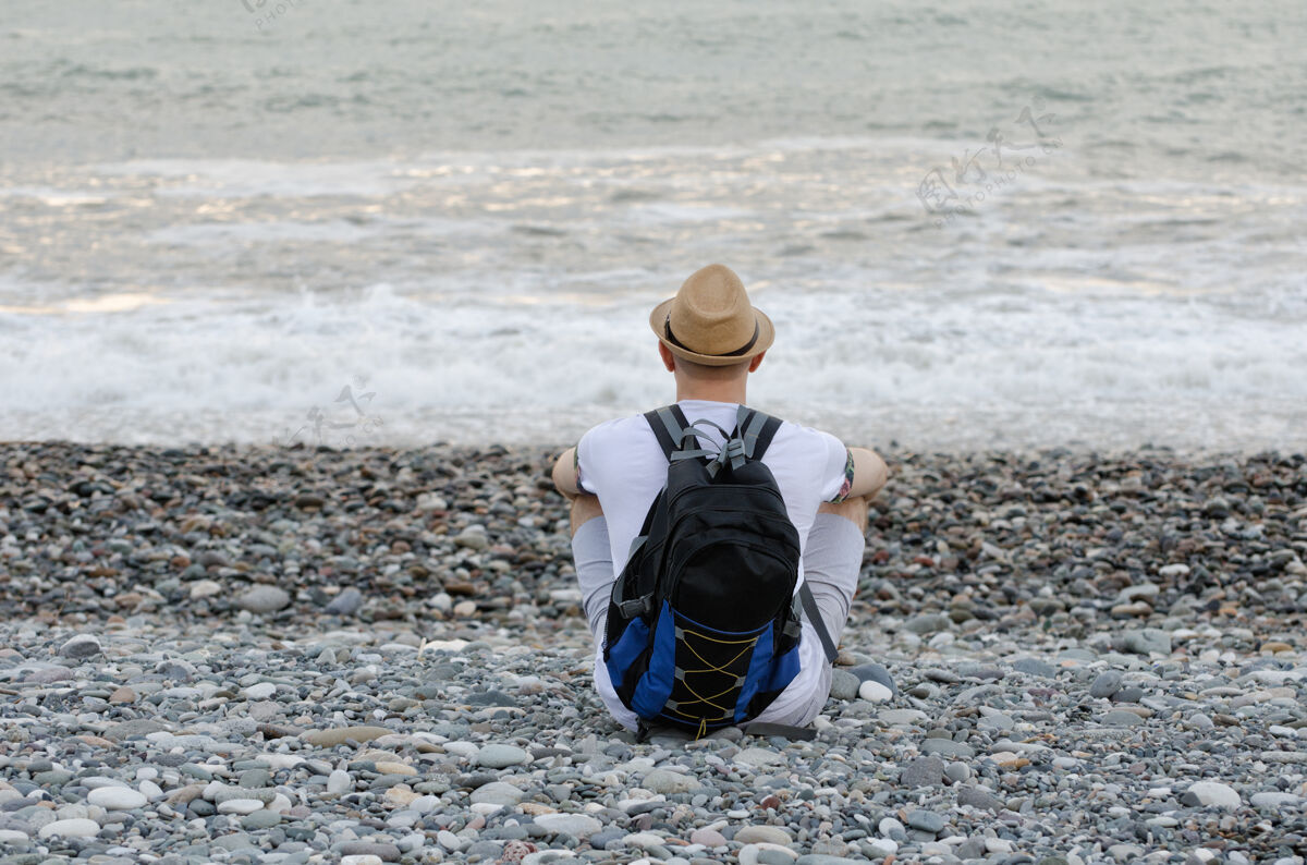 天空一个戴着帽子背着背包的年轻人正坐在沙滩上做背景度假单身背包