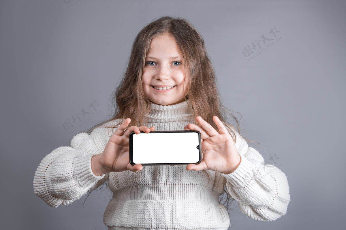 女性一个年轻漂亮的小女孩 金发飘逸的长发 穿着白色毛衣 在灰色的工作室里展示了一部白色空白屏幕的手机背景.地点为了文本.复制空间显示女孩复制
