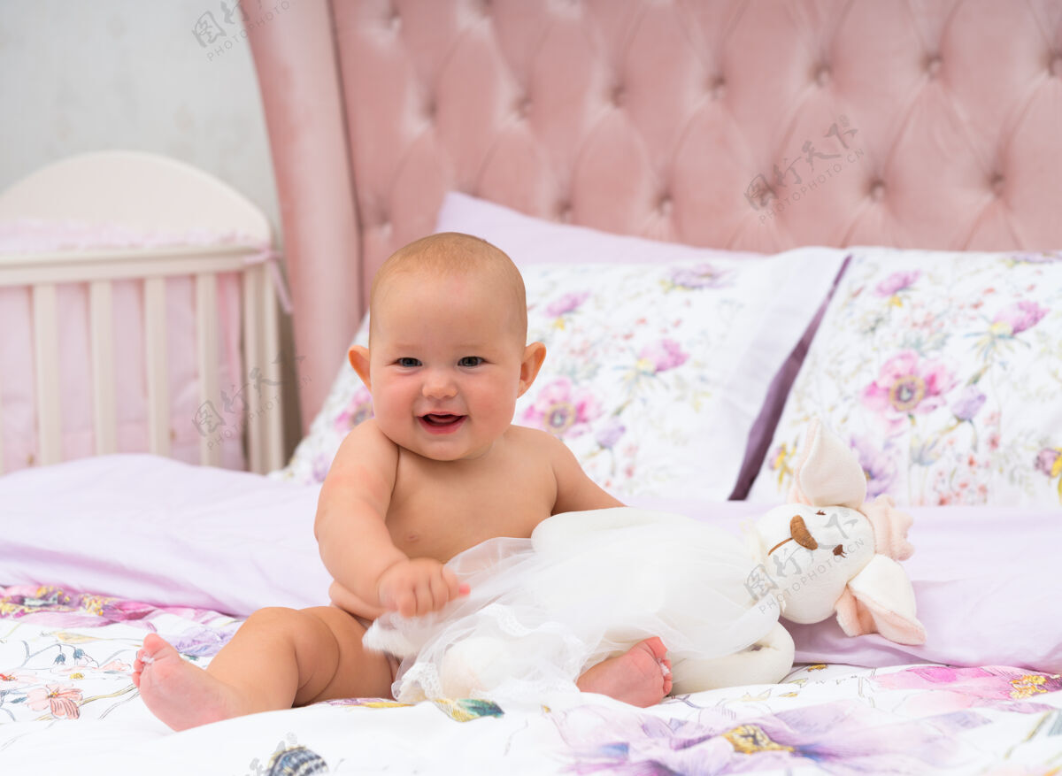 孩子快乐的胖乎乎的可爱小女婴坐在床上 抱着一个毛绒玩具 开心地对着镜头微笑肖像情感娱乐