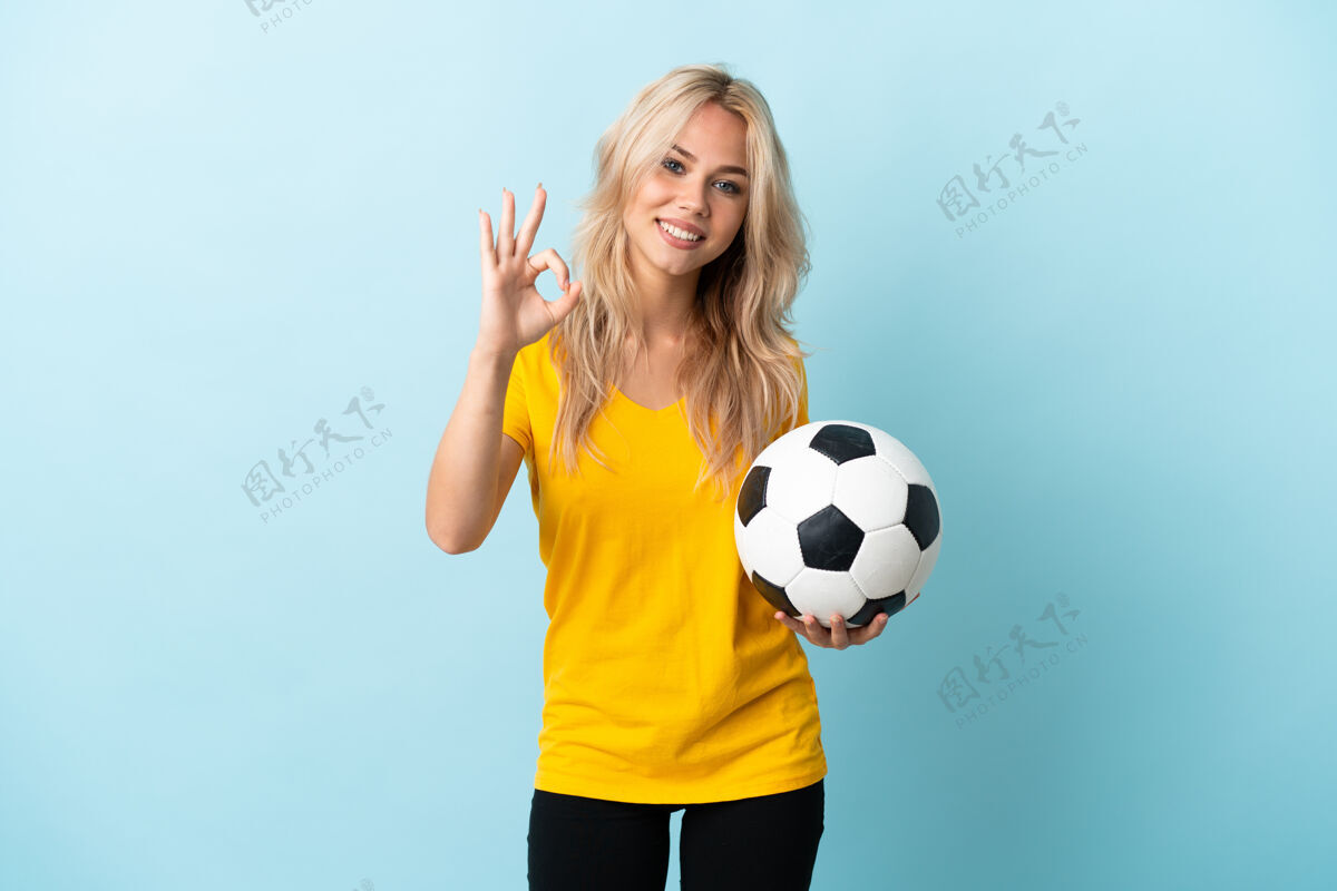 积极一个年轻的俄罗斯女人 拿着足球 在蓝色的地板上做着“ok”的手势女性手势Ok