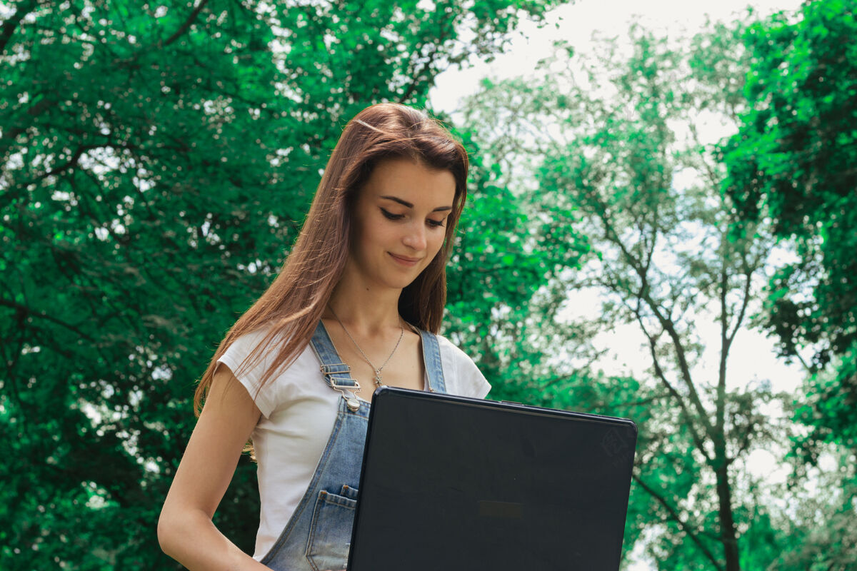 笔记本电脑美丽的年轻女士近距离地看着树丛中的一个黑色大笔记本学习服务漂亮