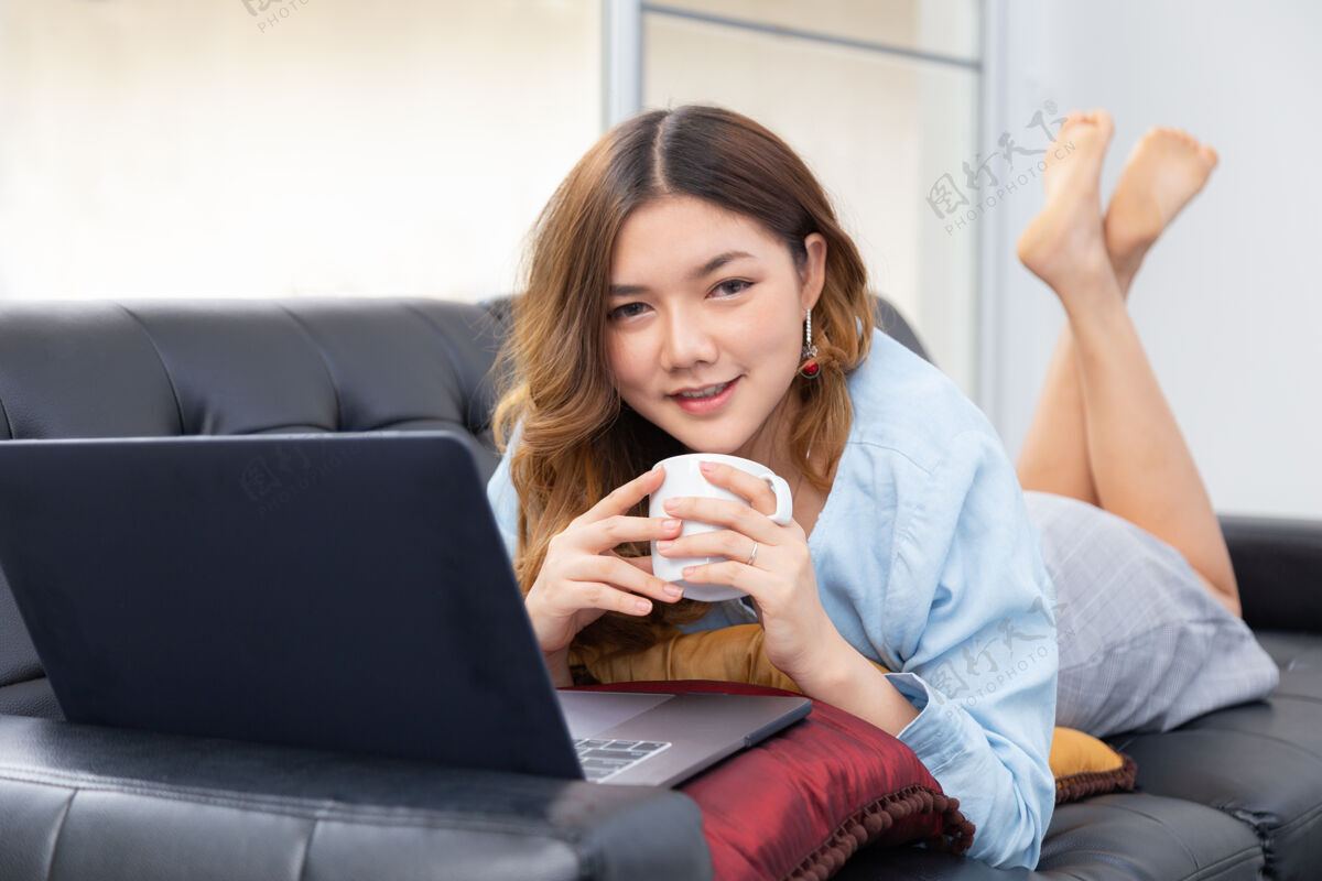 泰国美丽的亚洲年轻女子享受着在家的时光 一边躺在沙发上喝着热咖啡 一边用电脑工作爱好在线自拍