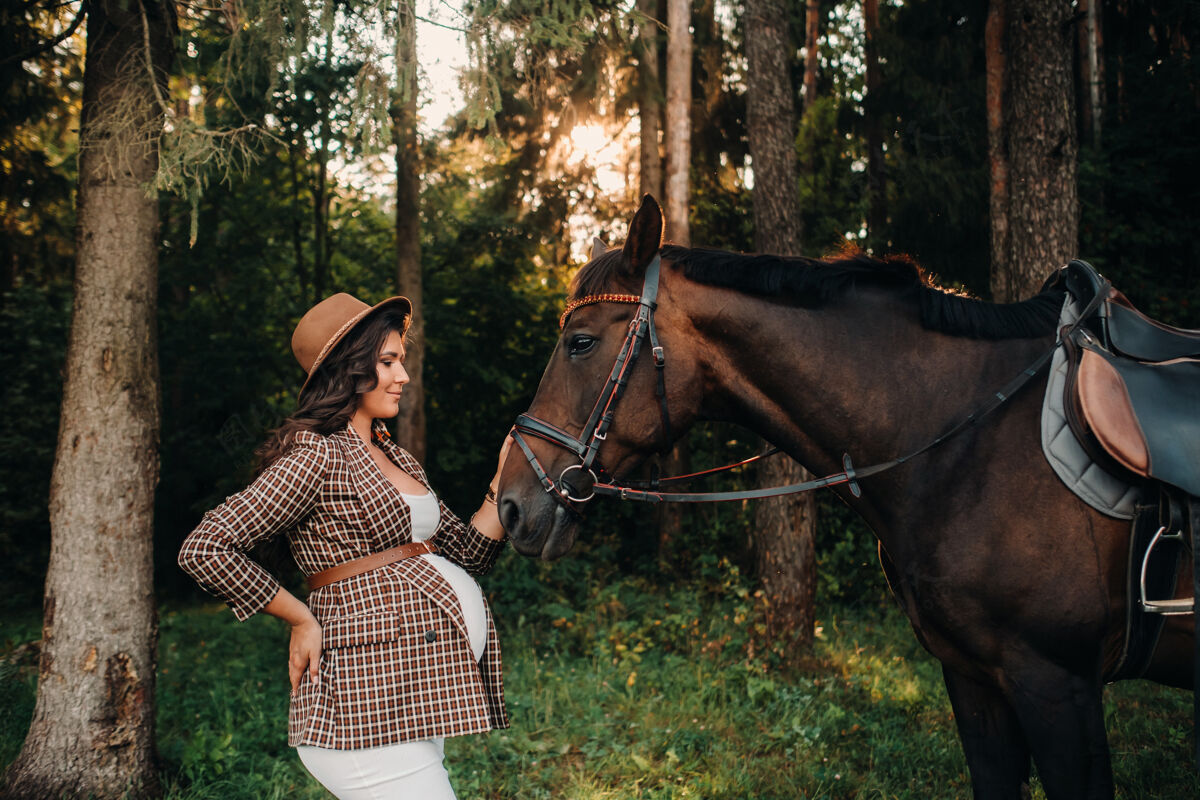 怀孕的母亲在大自然的森林里 一个戴着帽子的大肚子怀孕的女孩一个穿着白色裙子和棕色夹克的时髦孕妇 和马在一起肥肥父母腹部