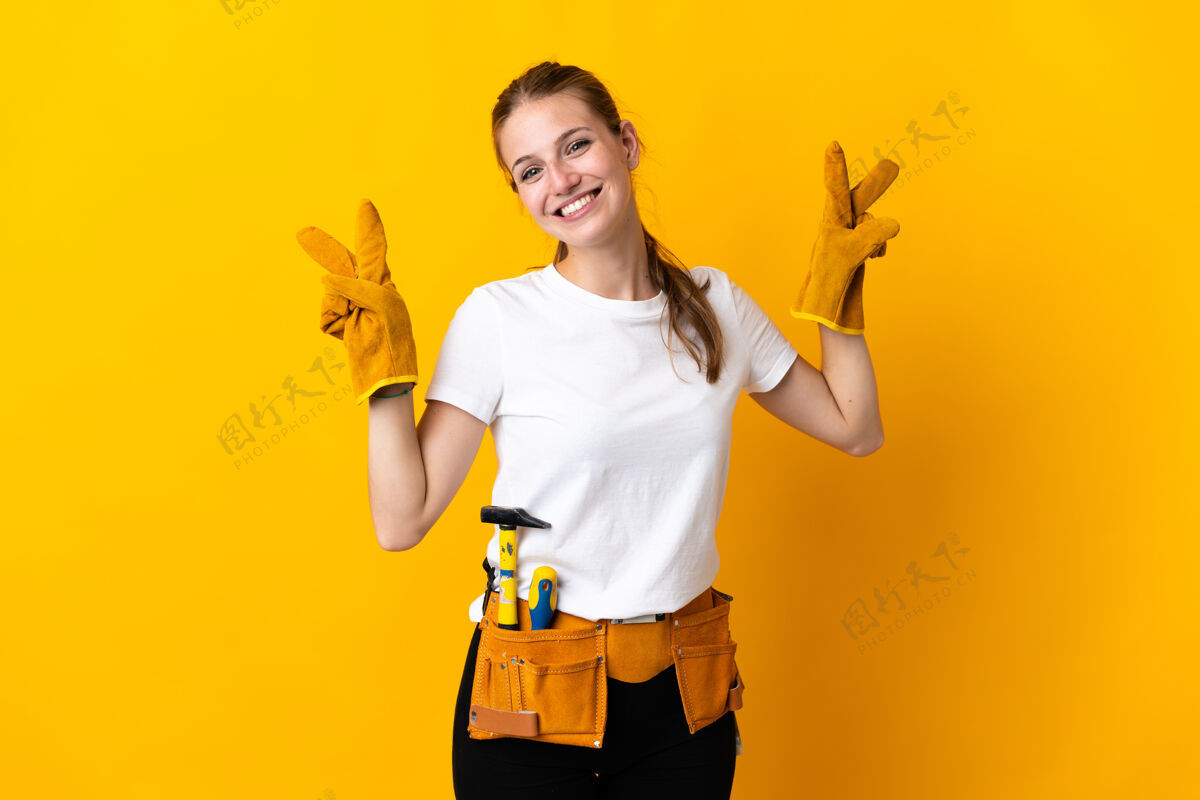 修理被隔离在黄色背景上的年轻女电工双手举着胜利的牌子工匠职业积极