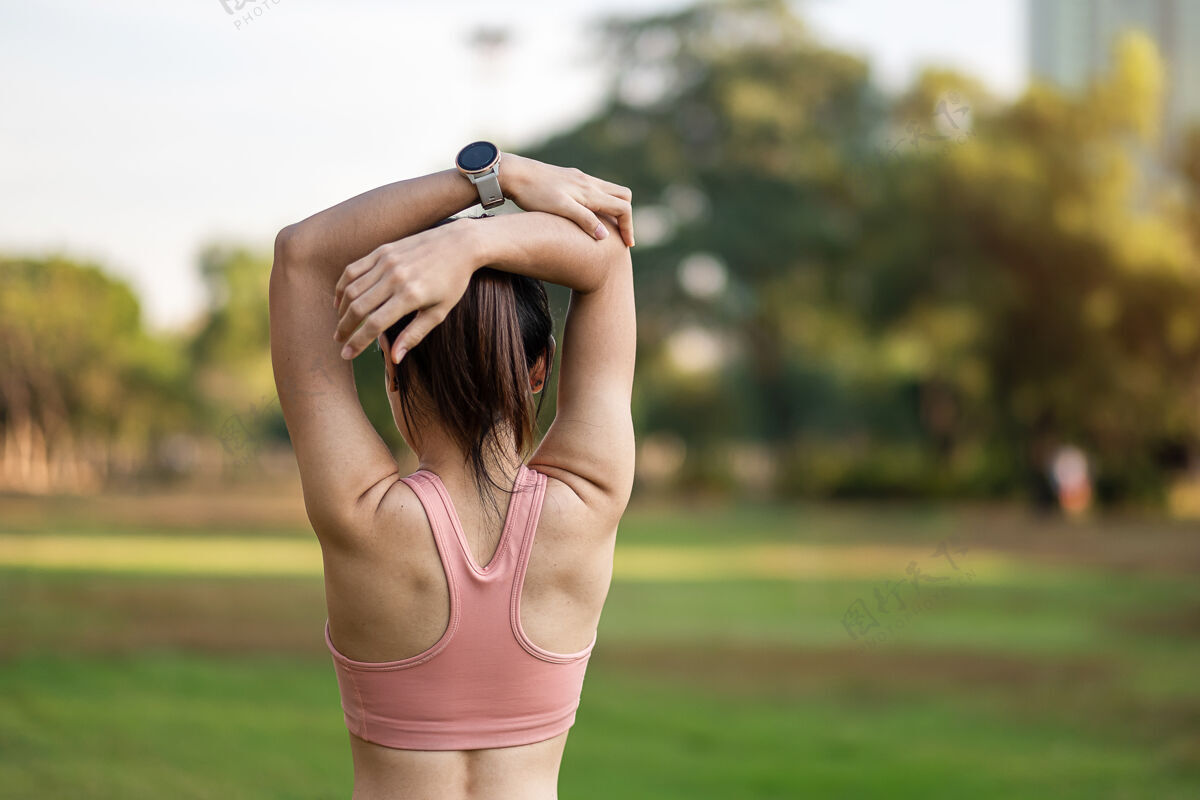 工作穿着粉色运动服的年轻成年女性在公园户外伸展肌肉 运动女性热身准备跑步和慢跑早上好健康 健身 运动和工作生活平衡概念锻炼肌肉身体