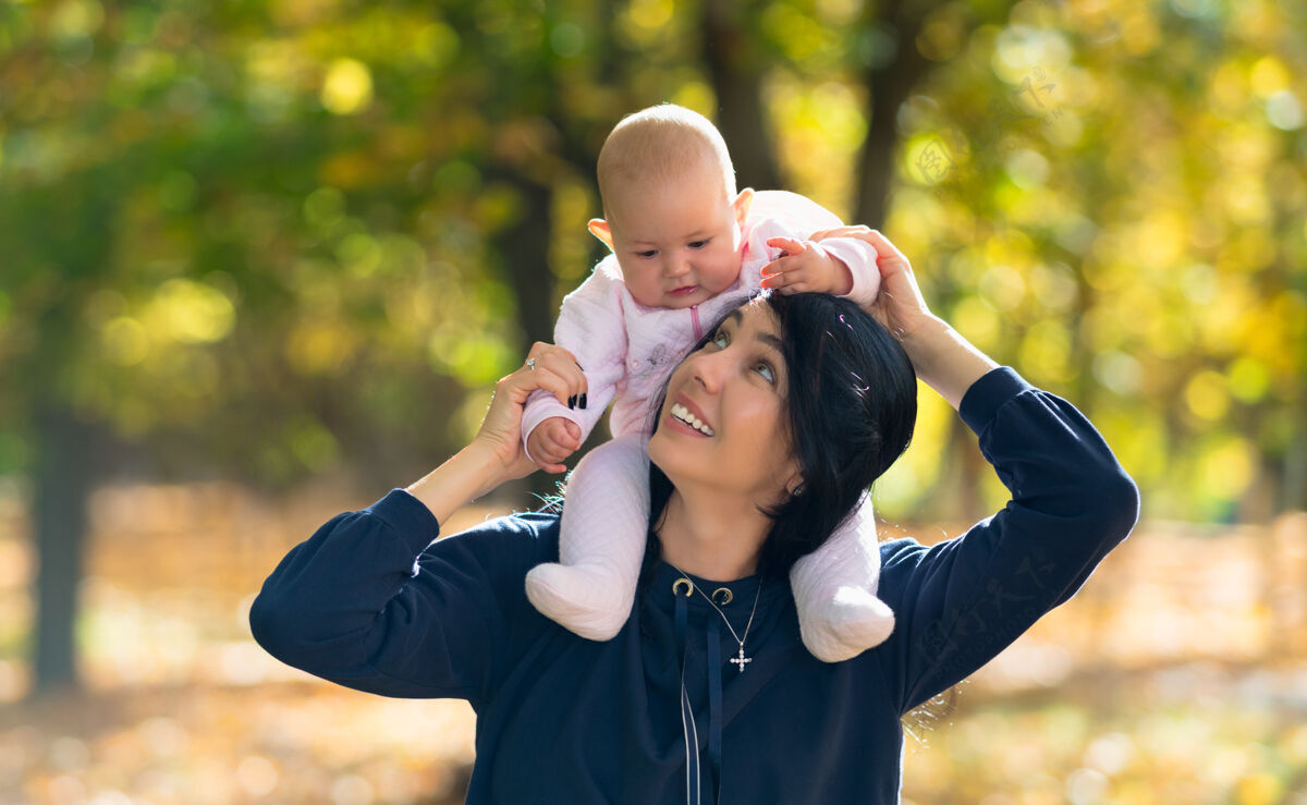 森林年轻的母亲和她的宝贝女儿在她的肩膀上微笑 因为她抬起头来 而在一个阳光灿烂多彩的秋季公园玩可爱可爱天真
