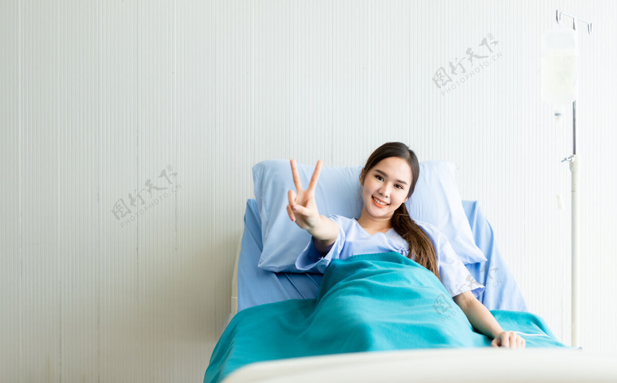 手亚洲女病人笑脸抬起两个手指在病房医院病床上与病魔搏斗孤独女人美丽