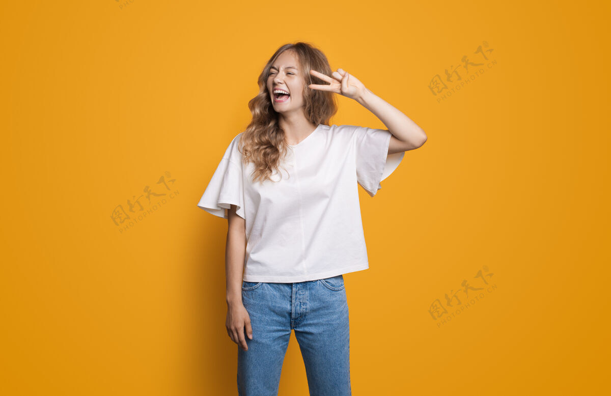 针织穿着白衬衫和牛仔裤的金发白种女人站在黄色的工作室墙上笑着 用手指做着和平的手势成功人青年