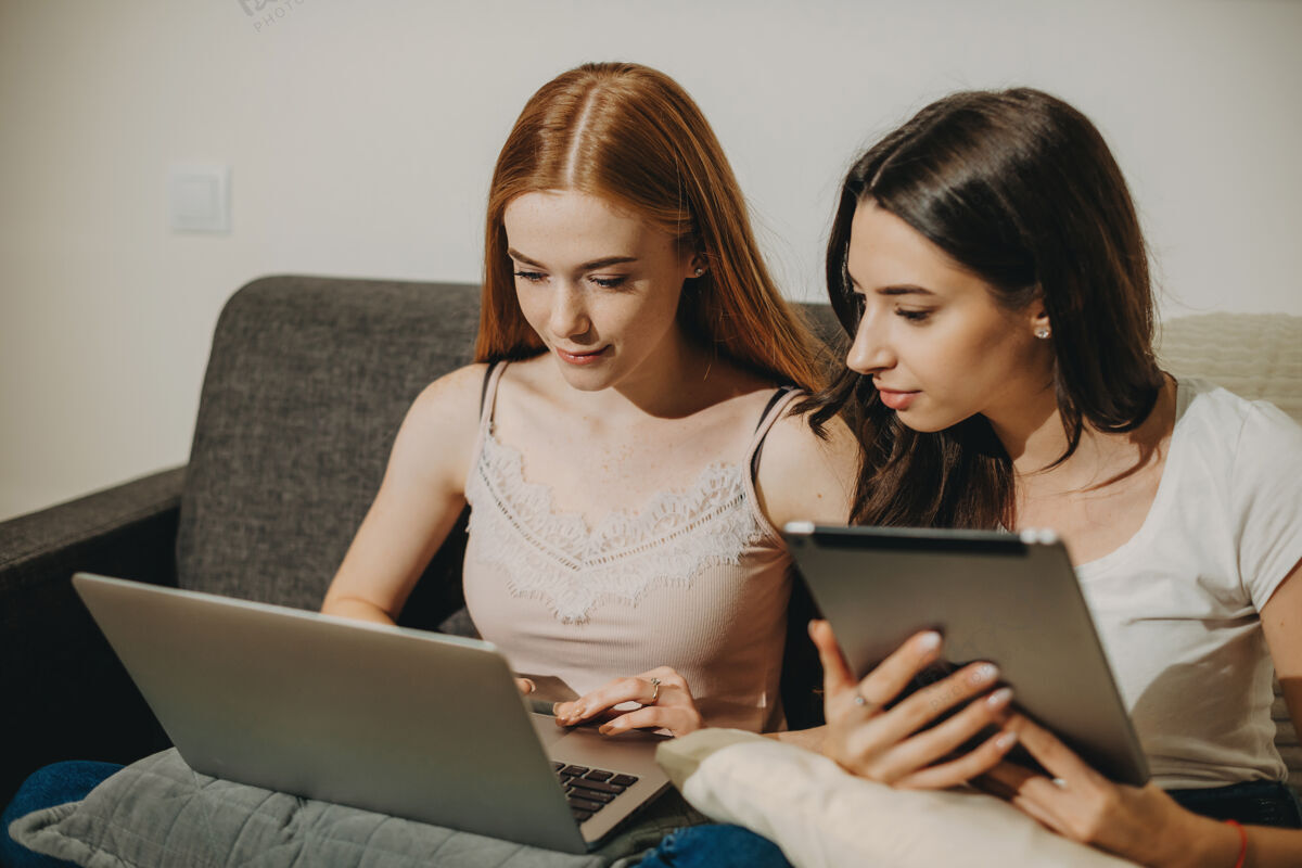 年轻年轻的红发女子看着笔记本电脑屏幕 而她的女朋友正拿着一个平板电脑坐在家里的沙发上平板电脑享受小玩意
