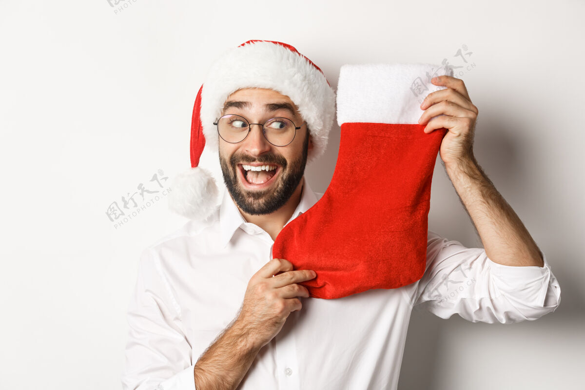 男朋友特写的快乐男人庆祝圣诞节 收到礼物在圣诞节袜子和期待兴奋 戴圣诞帽和眼镜 白色背景时尚帽子圣诞节