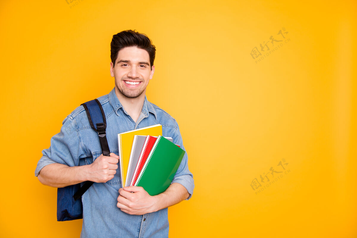 深色照片中 一个快乐积极的男人在空旷的空间附近拿着五颜六色的记事本 背着背包 隔着一堵亮色的墙男人年轻学习